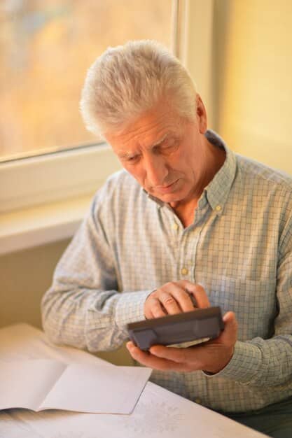 A senior man reviewing his budget on a tablet, with a calculator and various bills spread out on the table. He appears focused and organized.