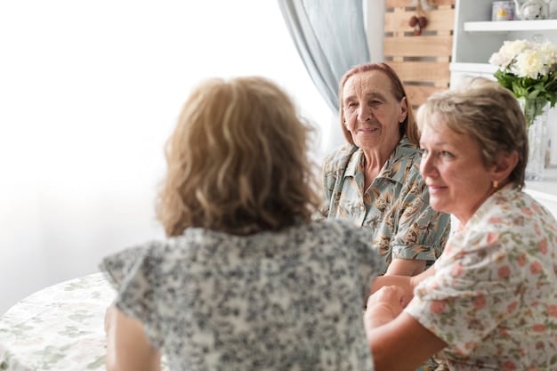 A senior woman is smiling and holding hands with her caregiver during a support group meeting. The setting is a community center with other caregivers in the background, creating a sense of camaraderie and mutual support. The focus is on the connection and emotional well-being of both the caregiver and the care recipient.